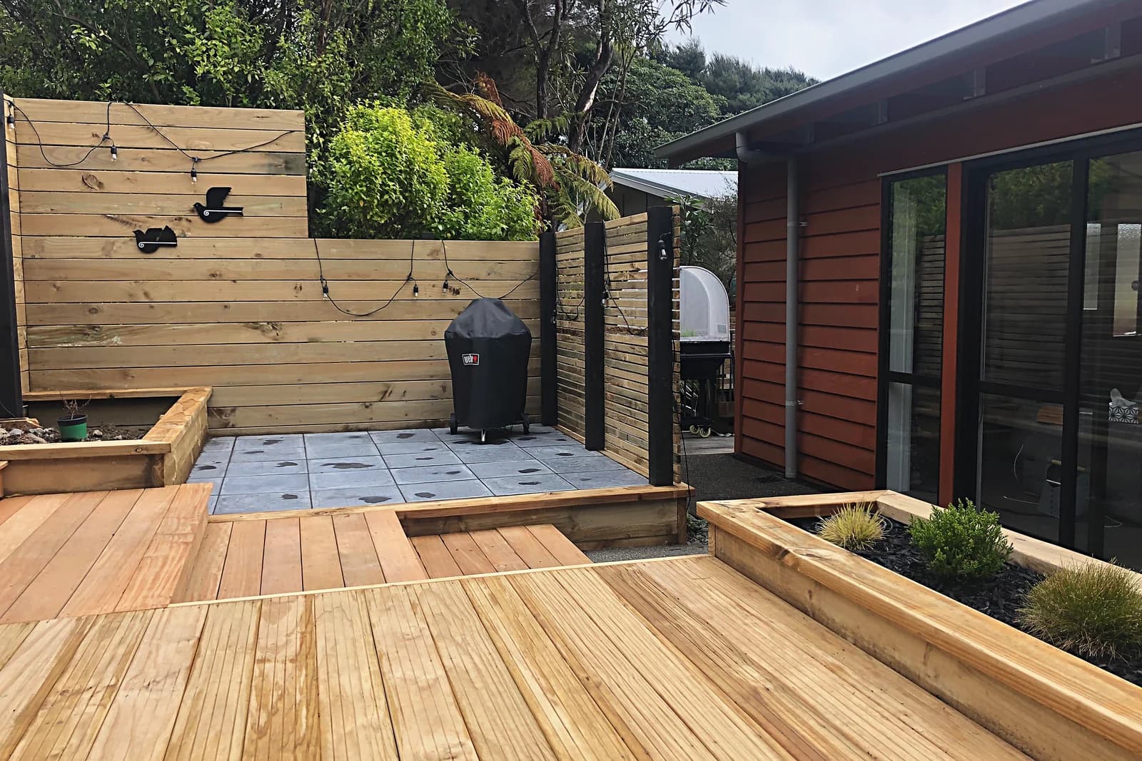 Timber deck stepping down to a paved corner with a slat screen and planter, Pukerua Bay